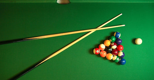 An aerial view of two cues lying crisscrossed on a pool table next to the pool balls set up in a triangle.